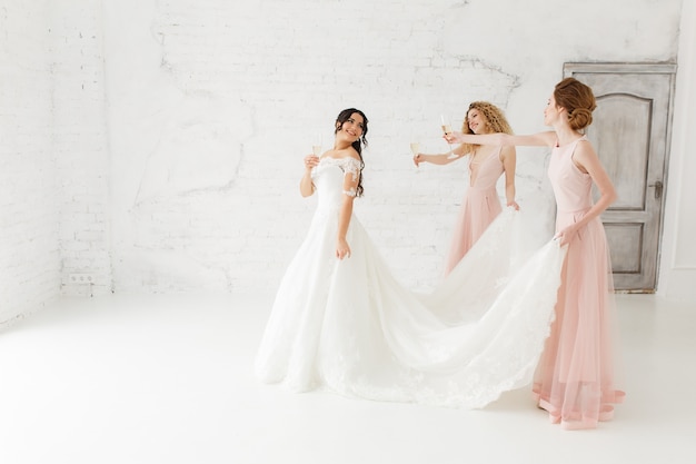 Happy beautiful bride and bridesmaid adjusting her wedding dress. Drinking champagne.