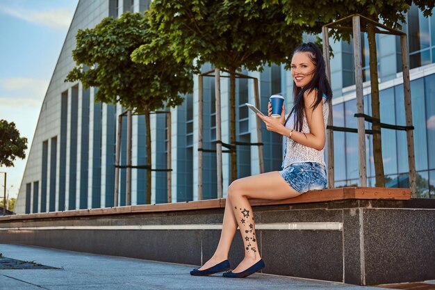 Happy beautiful brunette girl with tattoo on her leg wearing trendy clothes holds a takeaway coffee and a digital tablet while sitting on a bench against a skyscraper, looking at a camera.