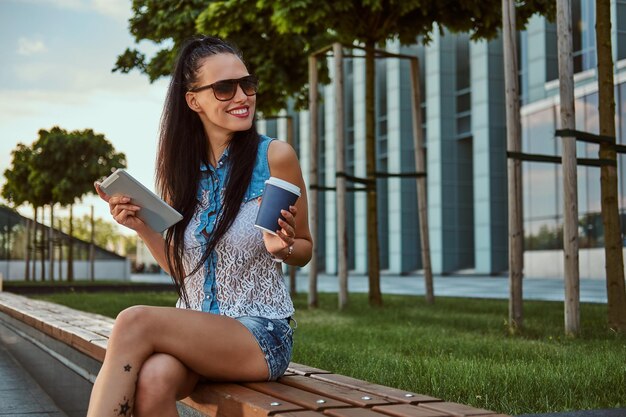 Happy beautiful brunette girl with tattoo on her leg wearing trendy clothes and sunglasses holds a takeaway coffee and a digital tablet while sitting on a bench against a skyscraper, looking away.