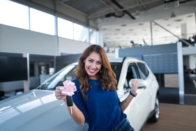 自動車販売店のショールームで新車の前に車のキーを保持している幸せな美しいブルネットの女性