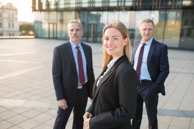幸せな美しいビジネス女性のオフィススーツを着て、屋外で立って、カメラ目線します。後ろに立っている男性のビジネス部門の同僚。ビジネスチームコンセプト