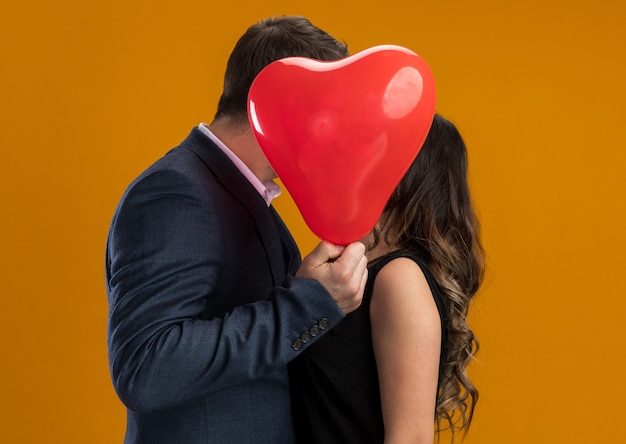 Felice e bella coppia che si bacia dietro un palloncino rosso a forma di cuore per celebrare il giorno di san valentino