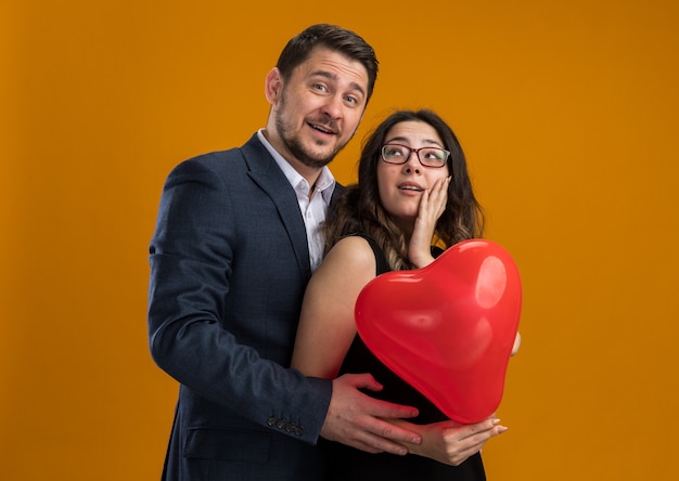 Happy and beautiful couple man and woman with red balloon in heart shape embracing celebrating valentines day over orange wall