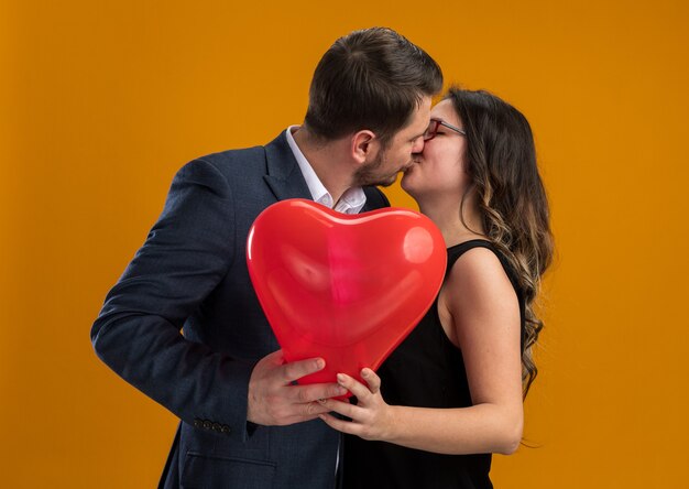 행복하고 아름다운 커플 남자와 여자 심장 모양의 빨간 풍선 포용과 오렌지 벽에 발렌타인 데이를 축하 키스