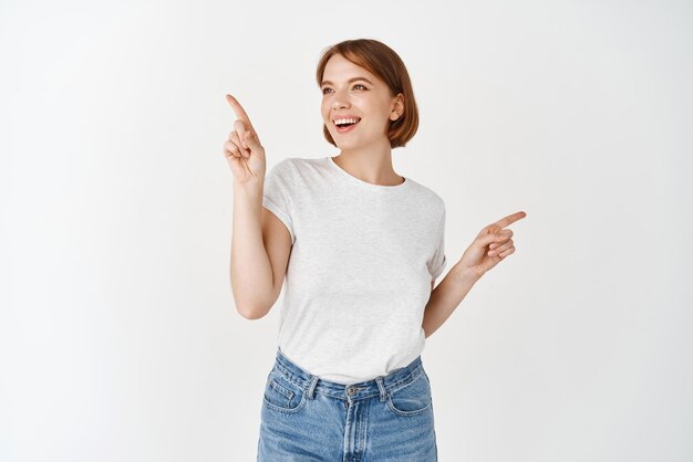 Happy beautiful female pointing sideways but looking left with cheerful smile making choice picking things on shopping standing against white background