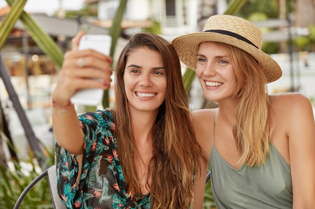 Happy beautiful females with broad smiles pose for selfie while sit together in cozy exotic cafeteria, use modern smart phone for making photo.
