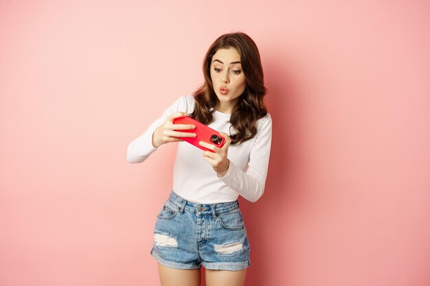Счастливая красивая девушка играет в мобильную видеоигру, держит смартфон горизонтально, смотрит на мобильный телефон с взволнованным лицом, розовый фон