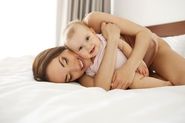 Happy beautiful mother in sleepwear lying on bed with her baby daughter embracing smiling.