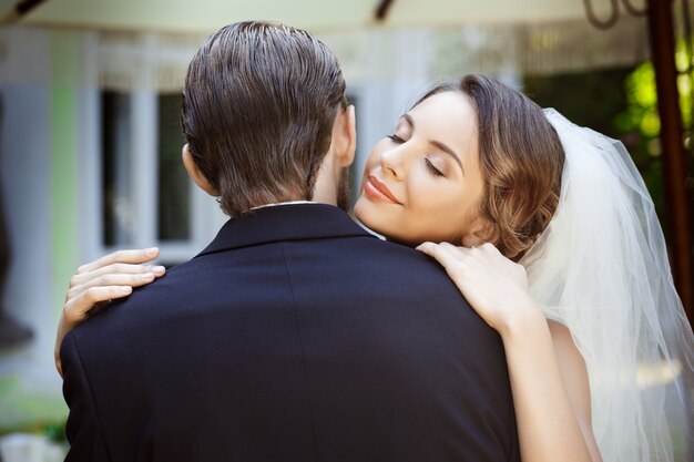 Happy beautiful newlyweds smiling, embracing in cafe outdoors. Eyes closed.