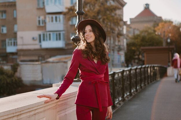 Happy beautiful stylish woman in purple suit walking in city street, spring summer autumn season fashion trend wearing hat, holding purse