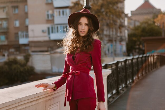 Happy beautiful stylish woman in purple suit walking in city street, spring summer autumn season fashion trend wearing hat, holding purse