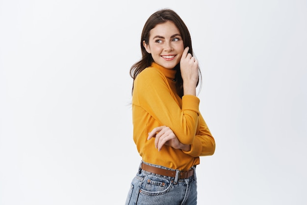 Foto gratuita felice bella donna in abiti casual, guardando lo spazio vuoto sul lato sinistro, sorridente e toccando il viso morbido e pulito, in piedi contro il muro bianco