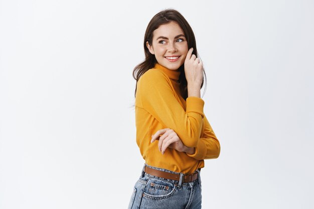 Happy beautiful woman in casual clothes, looking at empty space at left side, smiling and touching clean soft face, standing against white wall