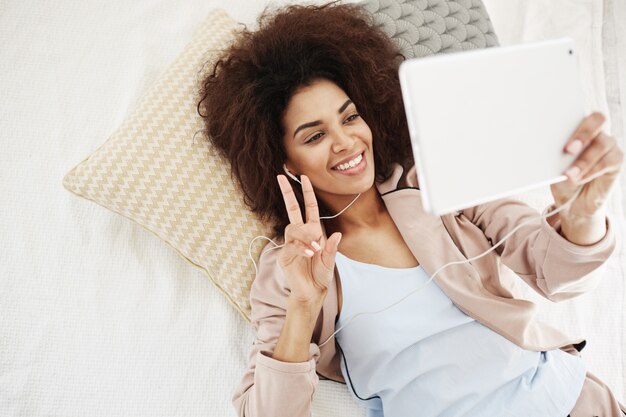 침대에 누워 태블릿에 평화 만들기 selfie 미소 헤드폰에서 행복 한 아름 다운 여자. 위에서.