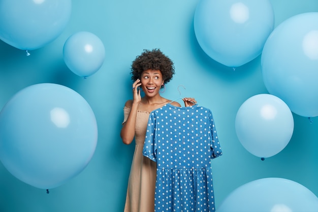 Happy beautiful woman holds blue polka dot dress on hanger, calls someone and uses her phone, prepares for special event, chooses outfit, poses around balloons. Clothing, wardrobe, fashion concept