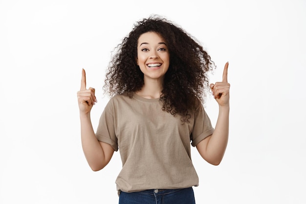 Happy beautiful woman pointing fingers up, smiling white teeth and showing advertisement on top, demonstrating empty space for banner, standing against studio background