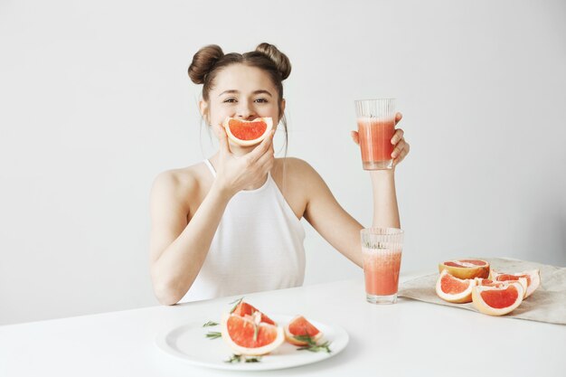 Счастливая красивая женщина сидя на таблице держа smoothie грейпфрута здорового вытрезвителя свежий над белой стеной.