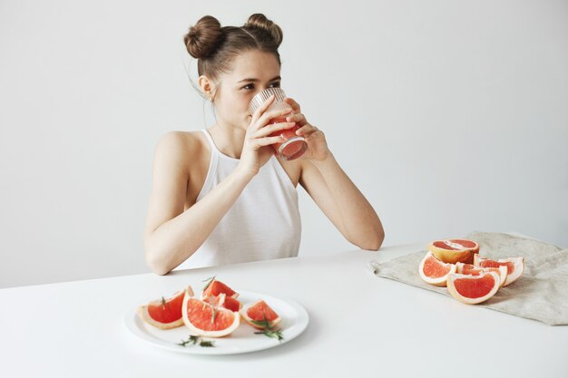 白い壁に健康的なデトックスフレッシュなグレープフルーツのスムージーを飲んでテーブルに座って笑って幸せな美しい女。