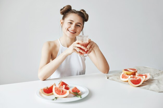白い壁に健康的なデトックスフレッシュなグレープフルーツのスムージーとガラスを保持しているテーブルに座って笑って幸せな美しい女。