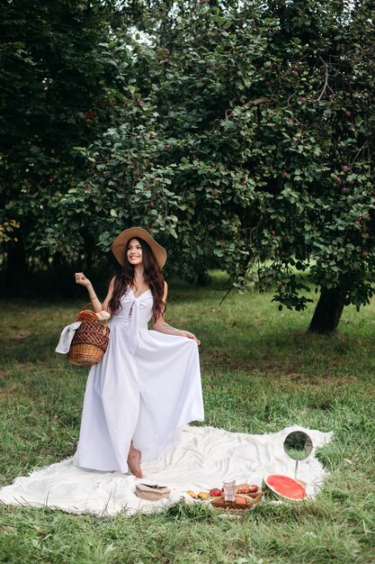 Happy beautiful woman wearing brimmed hat and white dress while standing and holding basket of bread in at summer park