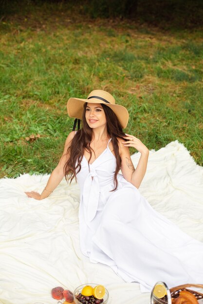 Happy beautiful woman wearing brimmed hat and white dress while standing and holding basket of bread in at summer park