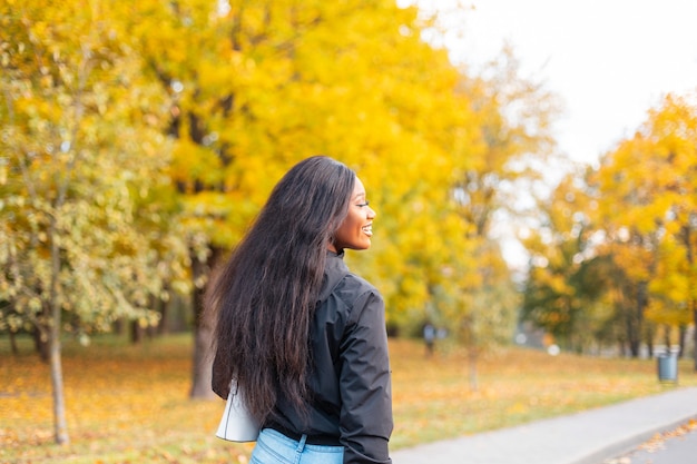Happy beautiful young african american woman with smile in trendy casual clothes walking in autumn park with bright colorful  yellow foliage