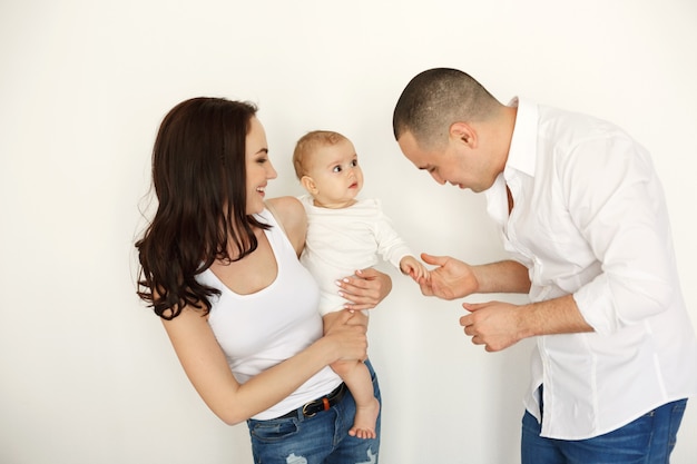 Happy beautiful young family with baby smiling embracing posing over white wall.