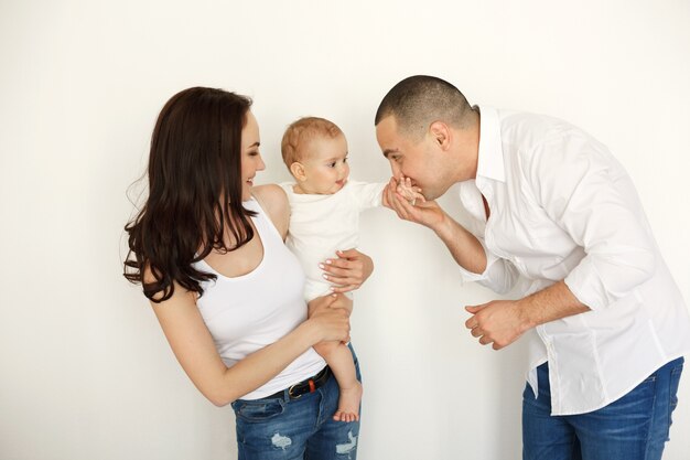 Happy beautiful young family with baby smiling embracing posing over white wall.