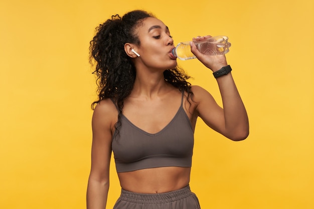 Happy beautiful young sportswoman listening to music using wireless earphones and drinking water from bottle isolated over yellow wall