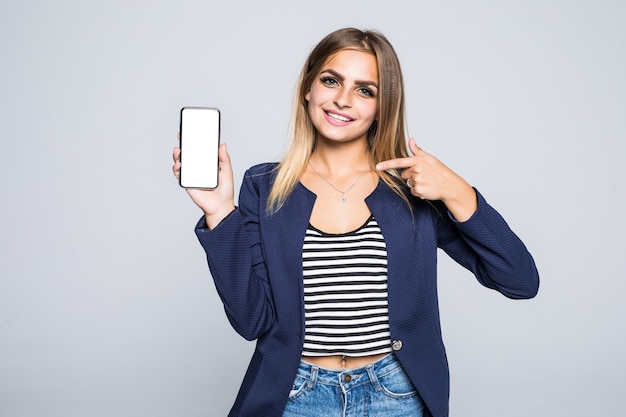 Happy beautiful young woman holding blank screen mobile phone and pointing finger over white wall