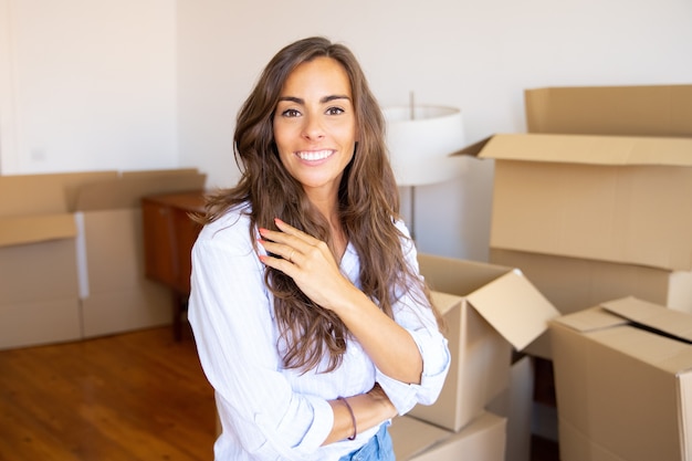 Happy beautiful young woman moving in new apartment, standing in front heap of opened carton boxes, looking at camera