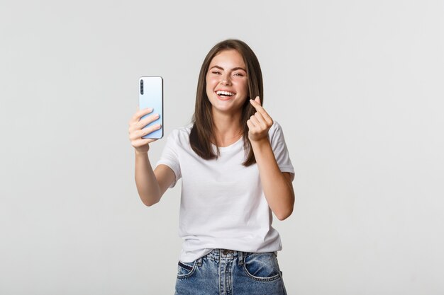 Счастливый красивая молодая женщина, показывая жест сердца и принимая selfie на смартфоне, беззаботно смеясь.