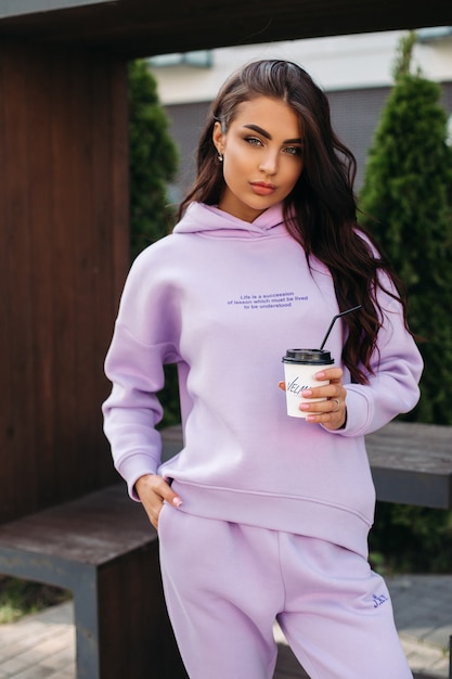 Happy beautiful young woman in sportswear standing on the street and holding coffee. Female fashion. City lifestyle