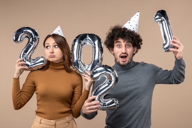 Happy bewildered uncertain young couple wear new year hat poses for camera Girl showing and and guy with and on gray