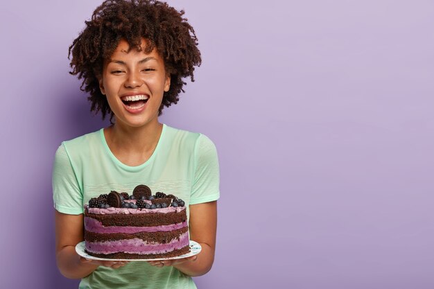 Happy birthday girl laughs joyfully, holds big tasty fruit cake, likes eating sweet food, improves mood with raising sugar in blood