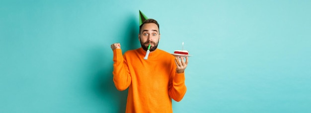 Free photo happy birthday guy celebrating wearing party hat blowing wistle and holding bday cake and doing fist