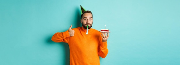 Free photo happy birthday guy celebrating wearing party hat blowing wistle and holding bday cake and showing th