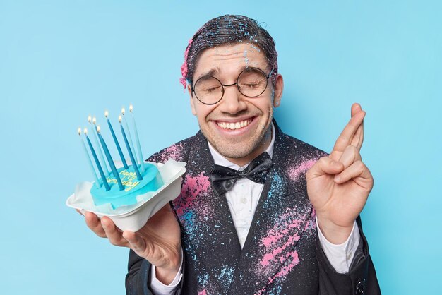 Happy birthday handsome man crosses fingers makes wish holds cake with burning candles dressed in festive clothes smiles broadly isolated over blue background Party time and celebration concept