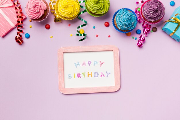 Happy birthday slate with colorful gems; streamers and muffins on pink background