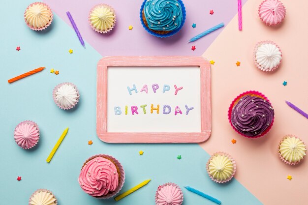 Happy birthday written on wooden frame surrounded with muffins; aalaw; sprinkles and candles on colored backdrop