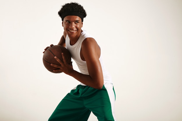 Счастливый черный баскетболист в зелено-белом костюме держит винтажный коричневый баскетбольный мяч, динамичная поза на белом