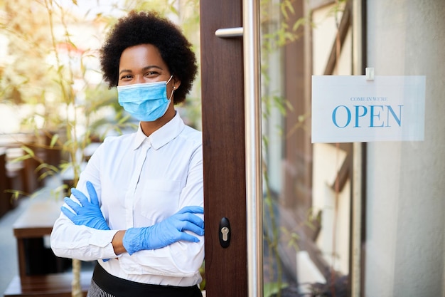 Happy black cafe owner with face mask standing at entrance door while reopening during virus epidemic