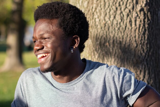 화창한 날 도시 공원에서 야외에서 시간을 보내는 행복한 흑인 남자. 회색 티셔츠를 입은 매력적인 아프리카계 미국인 남자가 옆을 바라보며 행복하게 웃고 있습니다. 근접 촬영입니다. 청소년, 라이프 스타일, 태도 개념입니다.