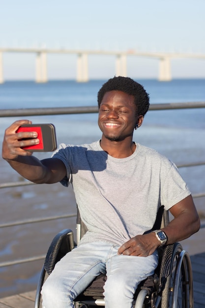 海岸で彼の携帯電話で自分撮りを取っている車椅子の幸せな黒人の男。携帯電話を持って笑っている若い男。バックグラウンドで橋。正面図。障害、現代の技術の概念。