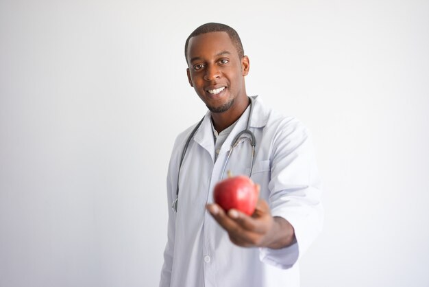 Happy black male doctor holding and offering red apple. 