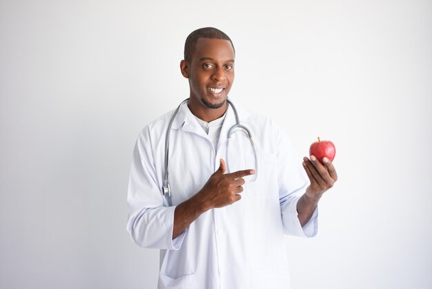 Happy black male doctor holding and pointing at apple. 