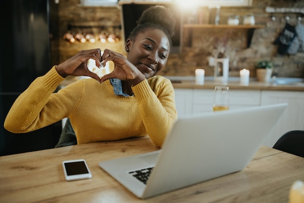 집에서 노트북으로 화상 통화를 하는 동안 누군가에게 심장 모양을 보여주는 행복한 흑인 여성