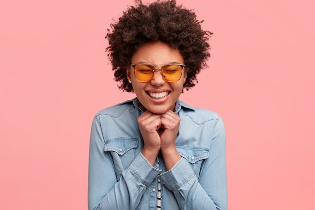 Happy black woman squints face from pleasure, smiles broadly, shows white teeth, keeps both hands under chin, wears yellow sunglasses, rejoices marriage proposal, isolated over pink wall