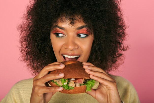 happy black woman with wavy hairs eating tasty cheeseburger on pink.