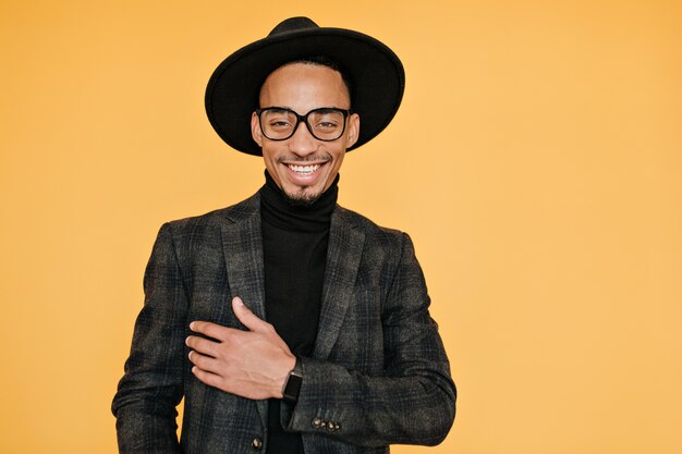 Happy black young man wears elegant dark suit posing with pleased smile. Indoor photo of relaxed mulatto male model in glasses enjoying photoshoot.